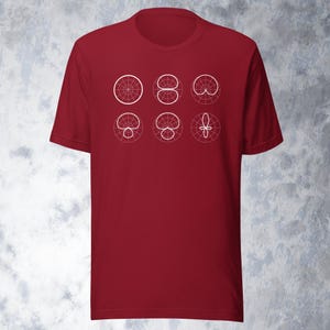 May include: A maroon short-sleeve t-shirt featuring six white geometric designs. The designs are circular and symmetrical, with varying patterns of lines and curves. The shirt is made of a soft material.