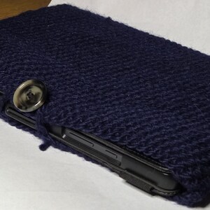 May include: A navy blue knitted tablet case with a large, round button closure. The case is made of a thick, textured yarn and appears to be handmade. The tablet is partially visible inside the case.
