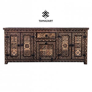 May include: A large, dark brown wooden cabinet with intricate geometric patterns carved into its surface. The cabinet features multiple doors, drawers, and decorative metal hardware. The brand name "TAMAZART" is displayed above the cabinet.