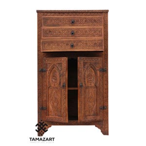Puede incluir: Un mueble de madera tallada a mano con intrincados patrones geométricos. El mueble tiene tres cajones y dos puertas que se abren para revelar un espacio de almacenamiento. El mueble es de color marrón cálido y tiene herrajes metálicos. El logotipo "TAMAZART" es visible.