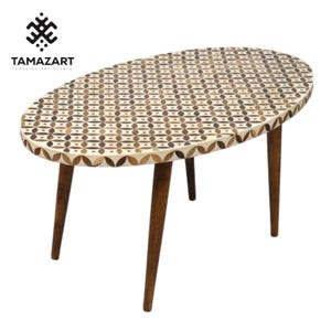 Puede incluir: Mesa de centro ovalada con una superficie estampada. La tapa de la mesa tiene un diseño repetitivo en tonos marrones y crema. La mesa tiene cuatro patas de madera marrón oscuro en ángulo. La marca "TAMAZART" es visible en la esquina superior izquierda.