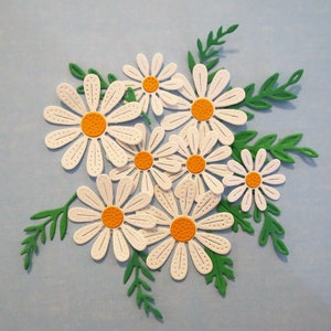 May include: A collection of seven white paper daisy die cuts with orange centers. The daisies are arranged in a cluster with green paper leaves around them.