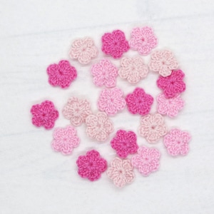 May include: A collection of small, five-petaled crochet flowers in various shades of pink, ranging from pale blush to vibrant magenta. Each individual flower is crafted with a central point and distinct, rounded petals, arranged in a loose cluster.