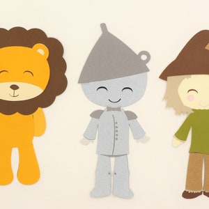 Puede incluir: Tres recortes de papel de personajes de El mago de Oz: un le&oacute;n con una melena marr&oacute;n y un cuerpo naranja, un hombre de hojalata con un cuerpo plateado y un sombrero gris, y un espantap&aacute;jaros con un sombrero marr&oacute;n, una camisa verde y pantalones marrones.