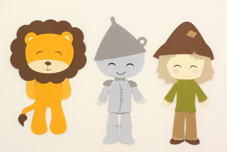 Puede incluir: Tres recortes de papel de personajes de El mago de Oz: un le&oacute;n con una melena marr&oacute;n y un cuerpo naranja, un hombre de hojalata con un cuerpo plateado y un sombrero gris, y un espantap&aacute;jaros con un sombrero marr&oacute;n, una camisa verde y pantalones marrones.