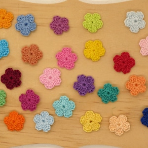 May include: A collection of colorful, crocheted flower appliques in various shades of blue, pink, orange, yellow, green, and purple. The flowers are arranged on a light brown wooden surface.