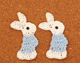 May include: Two crocheted bunny rabbit shapes with white and light blue yarn. The bunnies have black button eyes.
