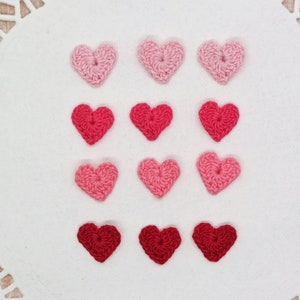 May include: Twelve crocheted hearts in various shades of pink and red, arranged in three rows of four hearts each.