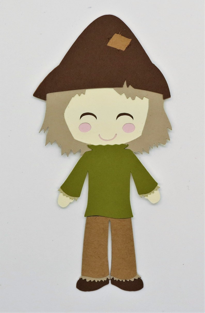 Puede incluir: Un recorte de papel marr&oacute;n y beige de un espantap&aacute;jaros con un sombrero marr&oacute;n, una camisa verde y pantalones marrones. El espantap&aacute;jaros tiene una cara sonriente con mejillas rosadas.