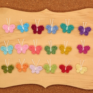 May include: A collection of 18 crocheted butterflies in various colors, including pink, blue, green, yellow, and purple, displayed on a wooden board.