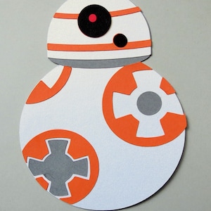 May include: A white and orange paper craft of a droid with a round body and a dome-shaped head. The droid has a black eye and a gray cross on its body.