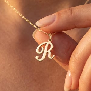 Könnte beinhalten: Eine goldfarbene Halskette mit einer feinen Kette und einem einzelnen Anhänger in Form des Großbuchstabens R in Schreibschrift. Der Anhänger ist an einem kleinen runden Verbindungsring befestigt, der an der dünnen, polierten Metallkette hängt.