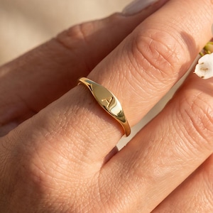 Puede incluir: Un anillo de sello de oro pulido que presenta una pequeña letra A grabada en la cara plana de forma ovalada. La banda es lisa y delgada, fabricada con un material metálico reflectante que capta la luz, creando un aspecto dorado brillante.