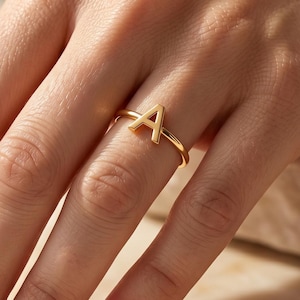 Könnte beinhalten: Ein goldfarbener Ring mit einem großen Buchstaben A als zentralem Blickfang. Der Ring ist dünn und kreisförmig gestaltet und besitzt eine polierte metallische Oberfläche, die das Licht reflektiert. Das A steht aufrecht auf dem Ring.