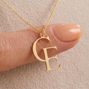 Könnte beinhalten: Eine goldfarbene Halskette mit einem Anhänger, der die überlappenden Großbuchstaben C und E zeigt. Der Anhänger hängt an einer zarten goldfarbenen Kette. Die Buchstaben sind in einer Serifenschrift mit polierter, reflektierender Metalloberfläche.