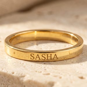 May include: A polished gold-tone band ring featuring the name SASHA engraved in a clean, sans-serif font on the outer surface. The ring has a smooth, reflective finish and a simple, uniform width throughout the entire circumference of the band.