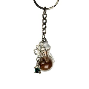 May include: A silver-toned keychain with a small glass vial filled with brown material. A silver clover charm and a small green gem dangle from the vial. The keychain has a round key ring and a chain.