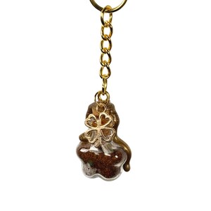 May include: A gold-coloured keyring with a small glass bottle charm. The bottle contains brown material and is topped with a gold-coloured four-leaf clover charm. The keyring is a decorative accessory.