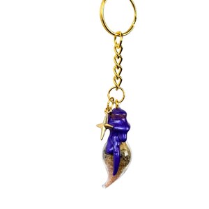 May include: A gold-coloured keyring with a chain and a charm. The charm is a small glass bottle filled with sand and herbs, sealed with a purple wax top. A gold star charm hangs next to the bottle.