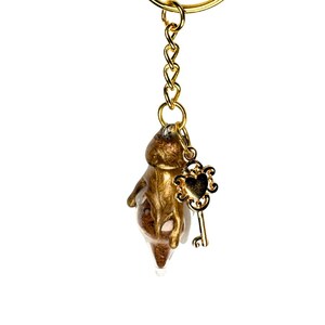 May include: A gold-colored keychain with a small, abstract figure encased in clear resin. The keychain features a gold ring, chain, and a decorative key charm with a heart-shaped design. The background is plain white.