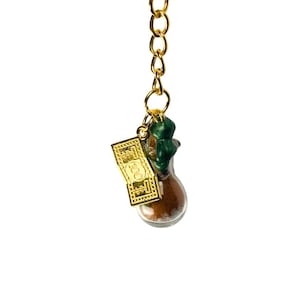 May include: A gold-coloured keyring featuring a small glass bottle filled with brown material. A gold-coloured $100 bill charm and a green stopper are attached to the bottle. The keyring is set against a white background.