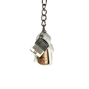 May include: A silver-colored keychain with a small laptop charm and a white figure attached to a cork. The keychain has a silver ring and chain. The cork has a brown and tan color.