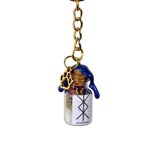 May include: A small glass bottle keychain filled with herbs and crystals, sealed with a cork and blue wax. A gold paw print charm and a gold keyring are attached. The bottle has a white label with the word "protection".