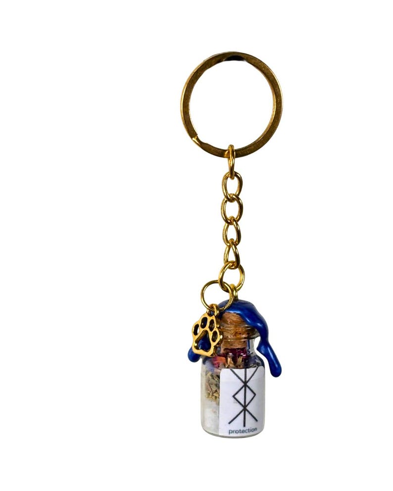 May include: A miniature glass bottle keyring filled with dried herbs and crystals, sealed with a cork and blue wax. A gold paw print charm and a gold keyring are attached. The bottle has a white label with the word "protection".