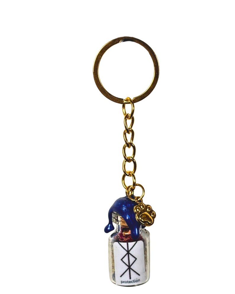 May include: A gold-coloured keyring with a small glass bottle charm. The bottle has a blue cap and a paw print charm. The bottle has a white label with a symbol and the word "protection".