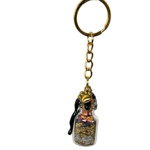 May include: A miniature glass bottle keyring filled with dried herbs and crystals. The bottle is sealed with black wax and a gold paw print charm. The keyring has a gold ring and chain.