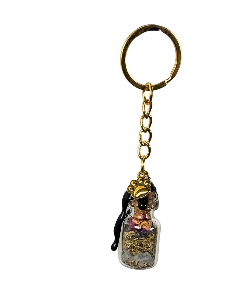 May include: A miniature glass bottle keyring filled with dried herbs and crystals. The bottle is sealed with black wax and a gold paw print charm. The keyring has a gold ring and chain.