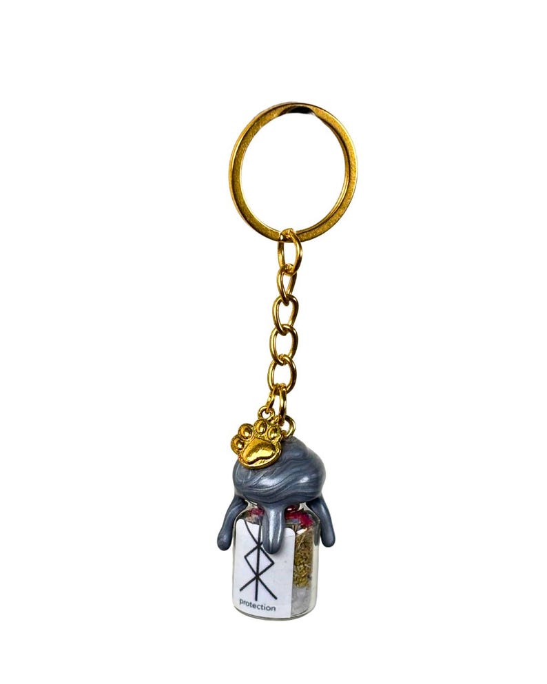 May include: A gold-coloured keyring with a small glass bottle pendant. The bottle is filled with herbs and has a grey, dripped-wax-like top. A gold paw print charm hangs above the bottle. The bottle has the word "Protection" and a symbol.