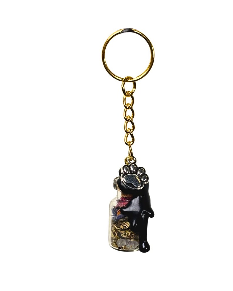 May include: A gold-coloured keyring with a small glass bottle pendant. The bottle contains dried flowers and small stones. A black paw print charm tops the bottle, with black liquid dripping down the side. The keyring is on a white background.