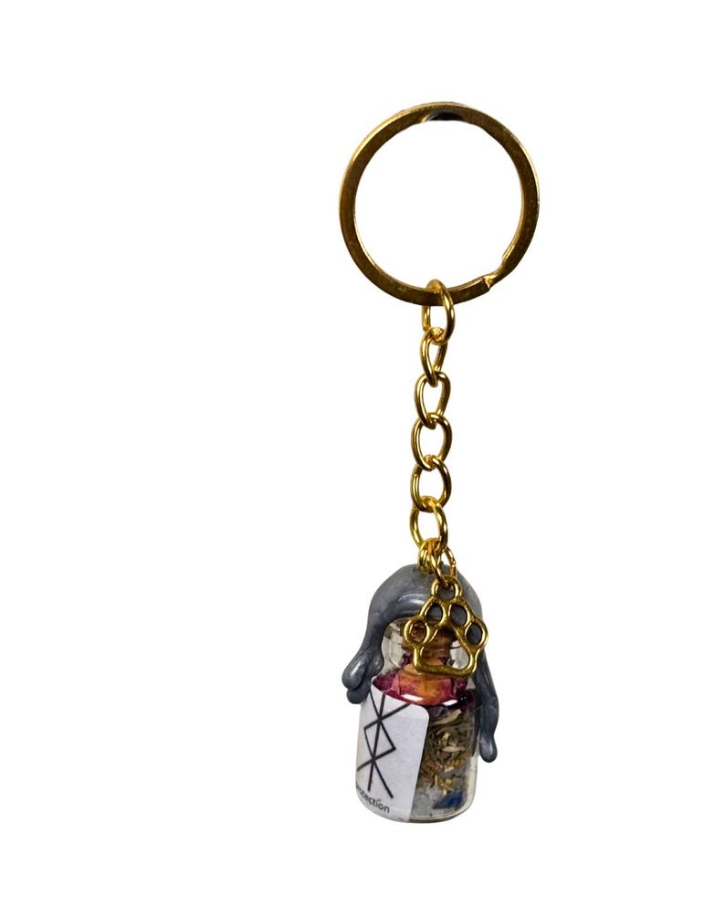 May include: A gold-coloured keyring with a small glass bottle pendant. The bottle contains dried herbs and flowers, sealed with grey wax and a gold paw charm. The bottle has a white label with black symbols.
