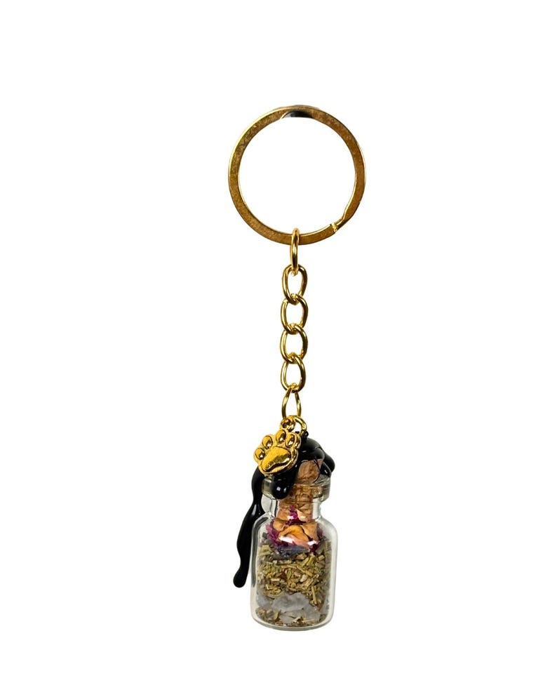 May include: A miniature glass bottle keyring filled with dried herbs and crystals. The bottle is sealed with a cork and adorned with a gold-coloured paw print charm. A gold chain and keyring complete the design.