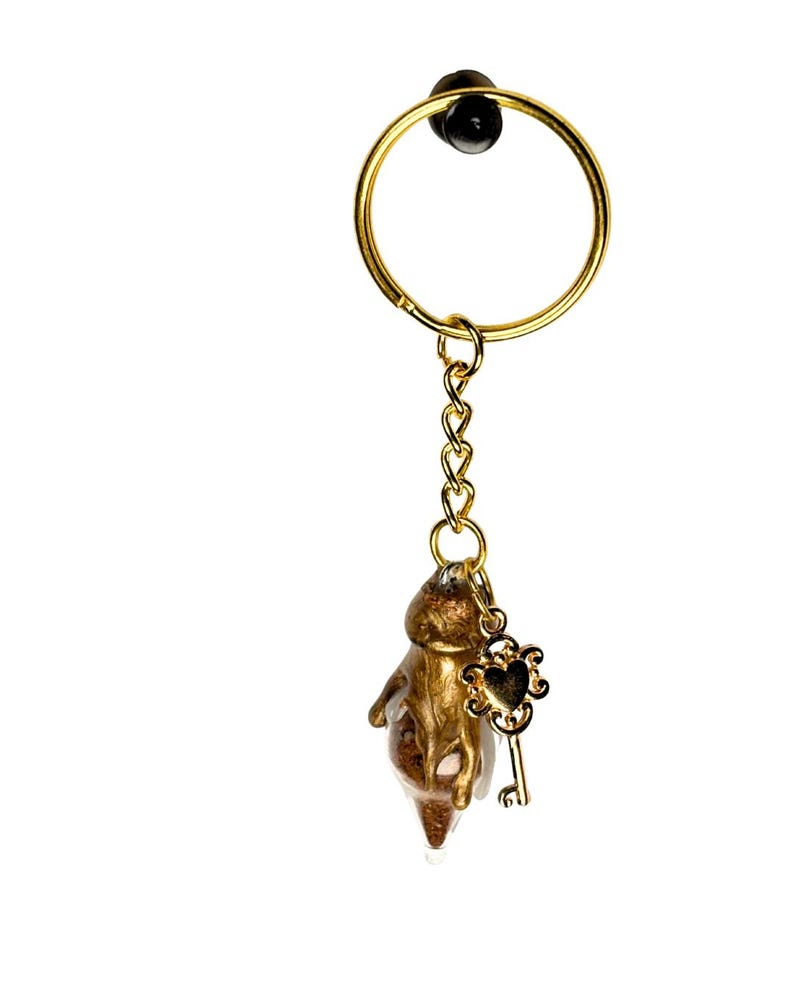 May include: A gold-colored keychain with a small, abstract figure encased in clear resin. The keychain features a gold ring, chain, and a decorative key charm with a heart-shaped design. The background is plain white.