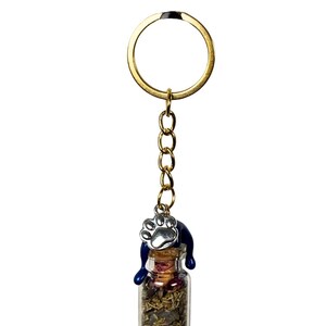 May include: A gold-coloured keyring with a small glass bottle filled with dried herbs and flowers. A blue cat-shaped charm sits atop the bottle, with a silver paw print charm hanging from the chain. The keyring is on a white background.