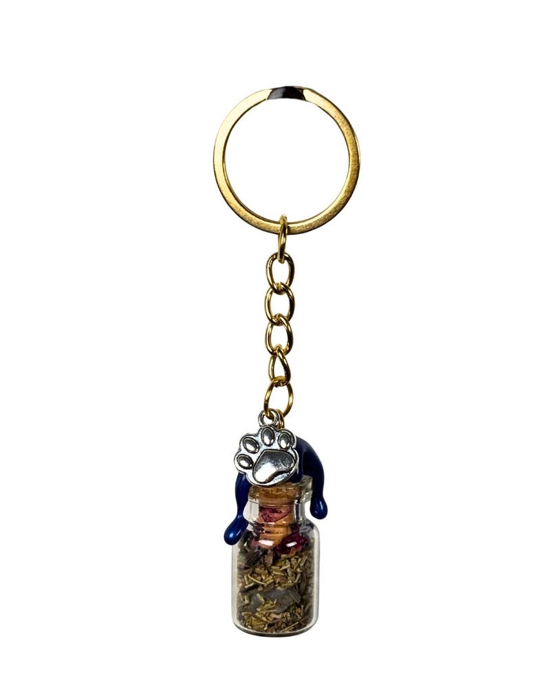 May include: A gold-coloured keyring with a small glass bottle filled with dried herbs and flowers. A blue cat-shaped charm sits atop the bottle, with a silver paw print charm hanging from the chain. The keyring is on a white background.