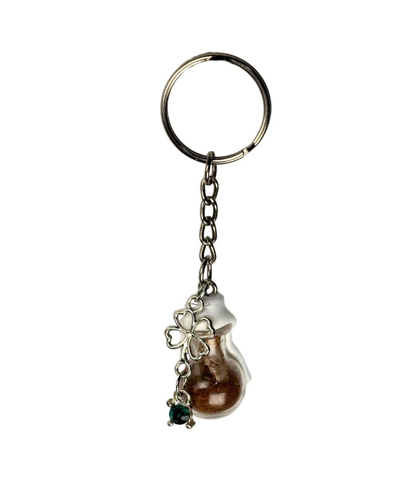 May include: A silver-toned keychain with a small glass vial filled with brown material. A silver clover charm and a small green gem dangle from the vial. The keychain has a round key ring and a chain.