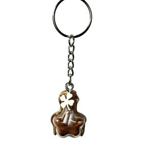 May include: A keychain with a silver ring and chain. The charm is a clear, rounded shape with a brown interior and a white clover on the front. The keychain is against a white background.