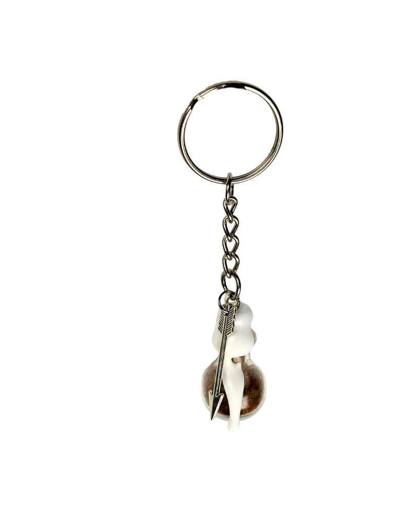 May include: A silver keychain with a small glass vial containing a brown substance and a white figurine pierced by a silver arrow. The keychain has a silver ring and chain.