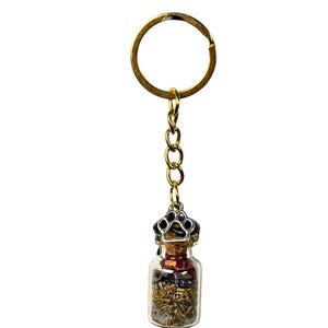 May include: A gold-coloured keyring with a small glass bottle pendant. The bottle is filled with dried herbs and flowers, and is topped with a silver-coloured paw print design. The keyring is a decorative accessory.