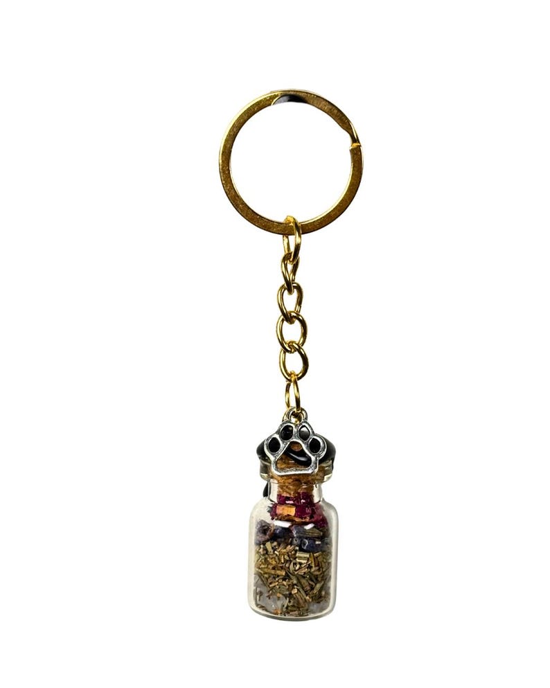 May include: A gold-coloured keyring with a small glass bottle pendant. The bottle is filled with dried herbs and flowers, and is topped with a silver-coloured paw print design. The keyring is a decorative accessory.