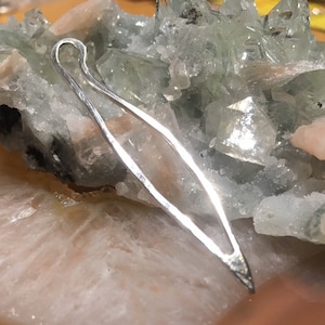 Peut inclure: Un pendentif en argent, ouvert aux deux extrémités, en forme de larme, avec une finition lisse et polie. Le pendentif repose sur un lit de cristaux verts et blancs.