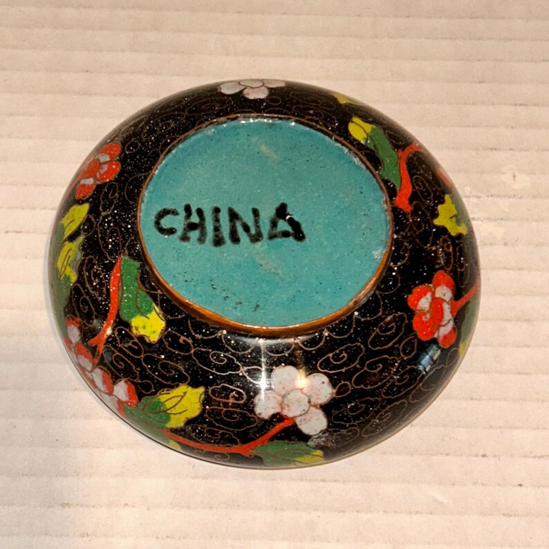 K&ouml;nnte beinhalten: Eine runde, schwarze Schmuckschatulle mit t&uuml;rkisfarbenem Innenraum. Die Au&szlig;enseite ist mit bunten Blumenmustern in Rot, Wei&szlig; und Gelb verziert. Das Wort "CHINA" ist in Schwarz auf dem t&uuml;rkisfarbenen Innenraum aufgedruckt.