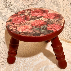 May include: A small, round wooden stool painted red. The top has a floral design with large pink and red roses. The stool has three turned legs, also painted red. The stool is suitable for decorative use, and measures approximately 25 cm in height.
