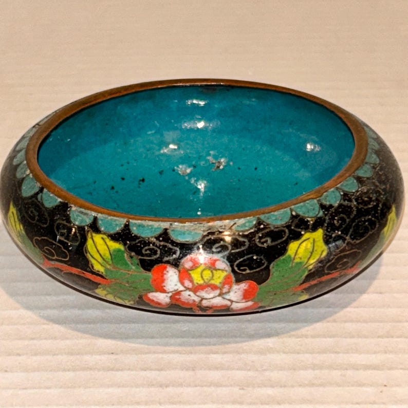 K&ouml;nnte beinhalten: Runde, dekorative Schale mit t&uuml;rkisfarbenem Innenraum und schwarzer Au&szlig;enseite. Das &Auml;u&szlig;ere zeigt ein Blumenmuster mit roten, gelben und gr&uuml;nen Akzenten. Der Rand ist bronzefarben.