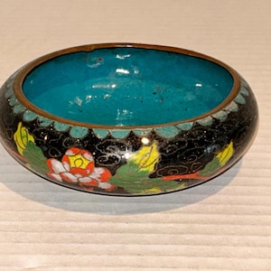 K&ouml;nnte beinhalten: Eine ovale, dekorative Schale mit schwarzer Au&szlig;enseite und t&uuml;rkisfarbenem Innenraum. Die Schale hat ein Blumenmuster mit roten, gelben und gr&uuml;nen Akzenten. Der Rand ist bronzefarben.