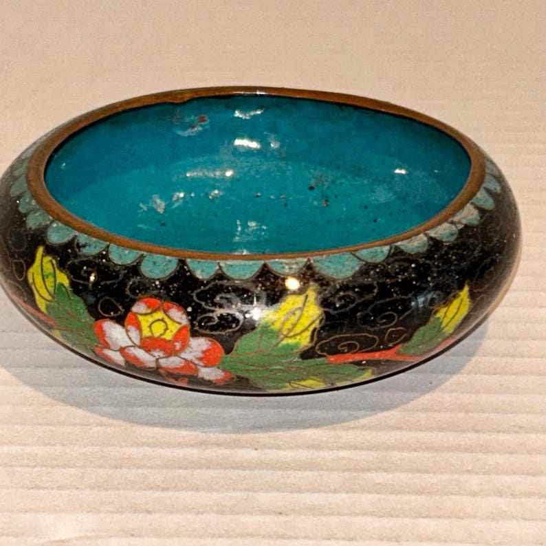 K&ouml;nnte beinhalten: Eine ovale, dekorative Schale mit schwarzer Au&szlig;enseite und t&uuml;rkisfarbenem Innenraum. Die Schale hat ein Blumenmuster mit roten, gelben und gr&uuml;nen Akzenten. Der Rand ist bronzefarben.