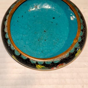 K&ouml;nnte beinhalten: Eine kleine, runde, antike Cloisonn&eacute;-Schale mit leuchtend t&uuml;rkisfarbenem Innenraum. Die Schale hat eine schwarze Au&szlig;enseite mit einem dekorativen Rand aus Blumen- und Blattmustern in verschiedenen Farben. Ein d&uuml;nnes, goldfarbenes Metallband umrandet den Rand.